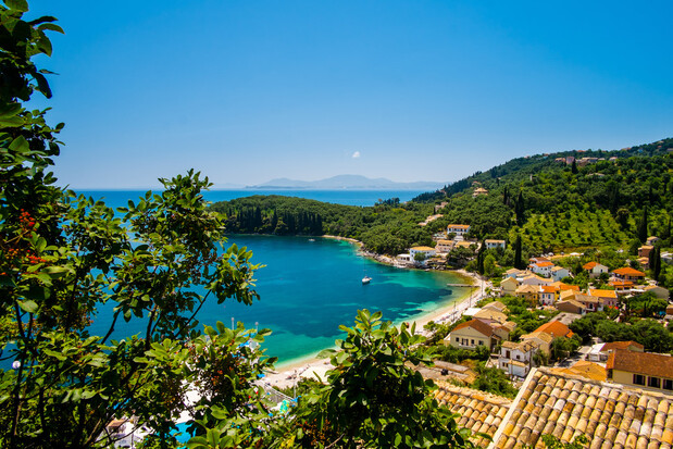 Coastal village nestled by a turquoise bay, surrounded by lush green hills and trees, with rooftops and boats dotting the idyllic scenery under a clear blue sky.